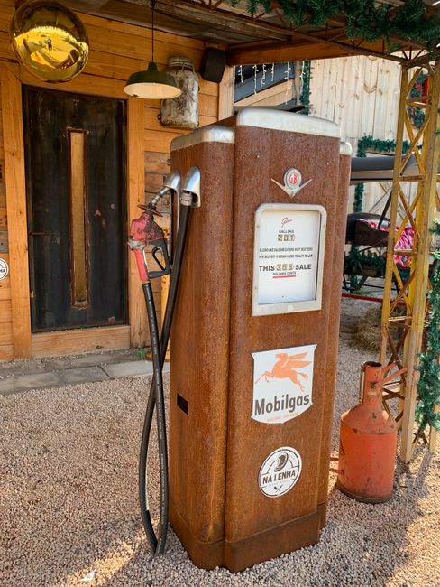 An outdoor fuel pump station with a rusted metal exterior, positioned on a gravel surface near a wooden building with a door and window. The pump has a white display area, with a Mobilgas logo featuring a red Pegasus above it. A fuel nozzle and hose are attached to the side of the pump, resting on a holder. To the right of the pump, there is an orange vintage gas cylinder standing upright. The environment includes decorative elements such as a green garland with flowers and a yellow metal framework, suggesting a rustic or outdoor setting. This scene visually relates to alternative waste handling or on-site fuel storage, possibly related to private or independent service options, aligning with the context of rubbish removal or similar services handled outside formal municipal systems.