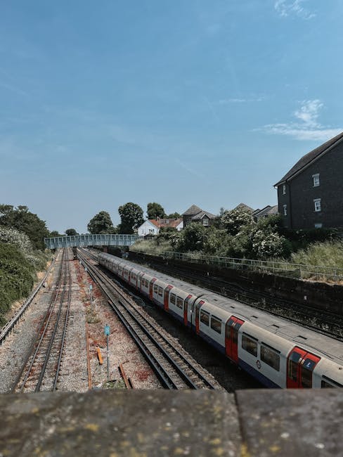 A photograph taken from an elevated position shows a section of railway tracks running through a residential area on a bright, clear day. The scene features multiple parallel train lines with gravel ballast and metal rails, some of which appear rusty. A modern commuter train with white and red exterior panels is parked on one track, partially visible along its length, with closed doors and windows reflecting daylight. On either side of the tracks, there are green shrubs and trees, with a few small white houses and dark brick residential buildings visible in the background, nestled among the foliage. The sky above is a vibrant blue with a few wispy clouds, suggesting calm weather. A white metal bridge spans across the tracks in the distance, connecting the residential area. The overall scene captures a typical commuter rail environment, with surroundings indicating an urban or suburban setting, and the photograph highlighting an alternative to traditional rubbish and waste collection through careful urban planning and infrastructure.