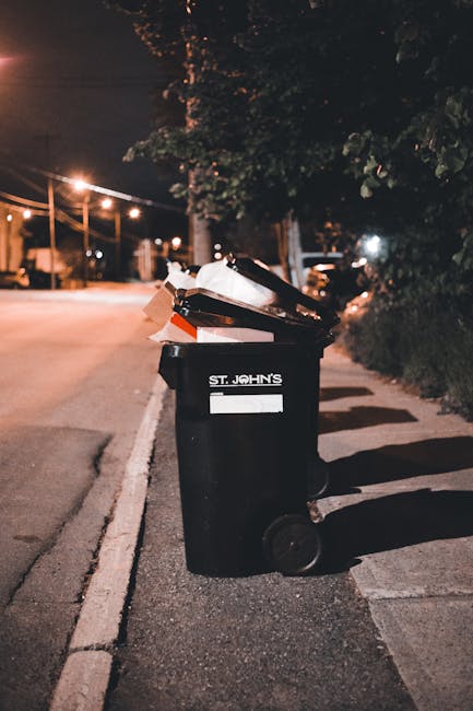 A black wheeled rubbish bin positioned on the curbside of a quiet street at night, with the label 'St. John's' visible on the front. The bin is overflowing with various discarded items, including flattened cardboard boxes, papers, and plastic waste, some of which are spilling over the top. The surrounding environment features a pavement with a white line marking the edge of the road, and a row of trees and bushes lining the sidewalk, partially illuminated by nearby streetlights. In the background, there are faint outlines of parked cars and streetlamps casting a warm glow over the scene. The overall scene is consistent with private waste collection or alternative rubbish removal services, highlighting the type of waste disposed of outside local authority collection times or in areas serviced by independent rubbish removal companies such as House Clearance Barnes, which offers professional rubbish clearance solutions in the SW13 area.