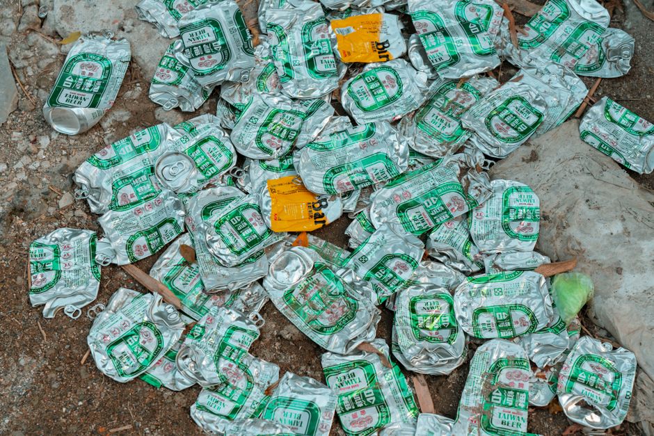A large scattering of crushed aluminum cans with green and white labels, including some orange-labeled items, lying on a dirt ground with small rocks, twigs, and leaves. The cans are irregularly piled, with some partially overlapping others, and appear to be emptied beverage containers, showing crumpled, dented textures. Surrounding the cans, there are pieces of discarded paper, a small plastic bag, and other debris, indicating a site where waste has been discarded haphazardly. The environment is outdoors, with natural lighting highlighting the metallic sheen and uneven surfaces of the cans, which suggest they might be remnants of a previous collection or casual littering. This scene reflects typical waste found in outdoor rubbish disposal areas, possibly associated with private or independent collection services like those offered by House Clearance Barnes, illustrating the need for rubbish removal or clearance services in such contexts.