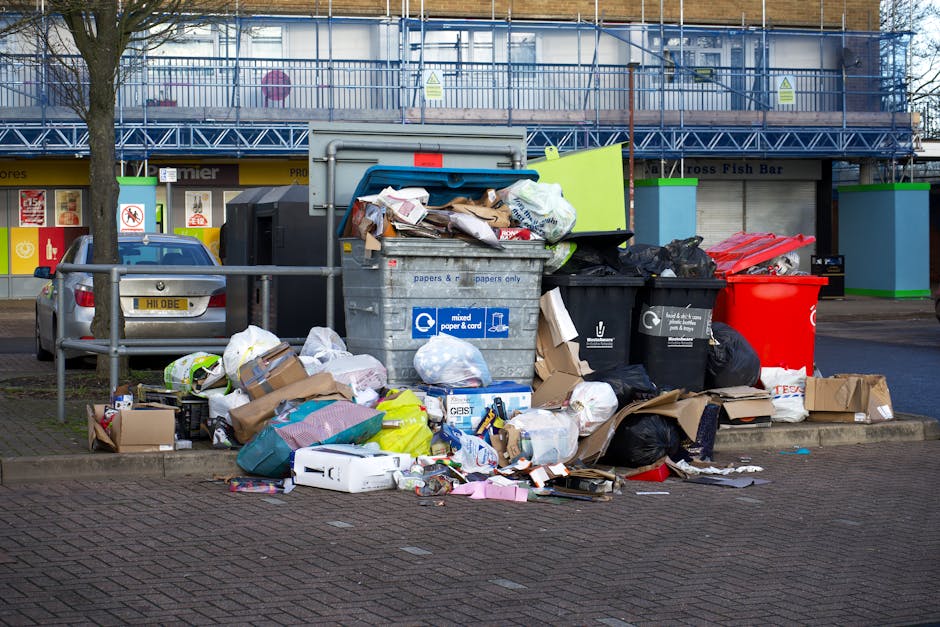 A collection of overflowing refuse bins and scattered rubbish situated on a paved urban pavement near a parking area. The scene features a large gray mixed paper and card bin with its lid open, revealing assorted flattened cardboard boxes, paper debris, and plastic packaging spilling over the edges. Several black plastic trash bags are piled against the bins, some partially torn, along with a bright red recycling bin also filled with waste. Surrounding the bins are various discarded items, including cardboard boxes, paper bags, plastic containers, and miscellaneous packaging, occupying most of the foreground. Behind the waste, part of a silver car with a visible license plate and parked on the street is seen, along with a protective fence and a building with storefronts and signage. The background includes a multi-storey building with metal scaffolding and safety netting, indicating ongoing maintenance or renovation. The scene is lit with natural daylight, emphasizing the cluttered state of the rubbish collection area, which appears neglected and ready for professional rubbish removal services like those provided by House Clearance Barnes, supporting private waste handling alternatives in the area.