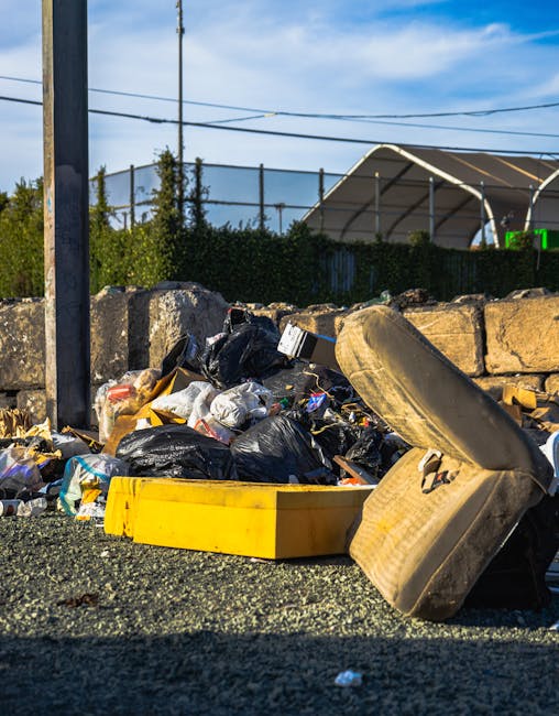 A collection of overflowing refuse bins and scattered rubbish situated on a paved urban pavement near a parking area. The scene features a large gray mixed paper and card bin with its lid open, revealing assorted flattened cardboard boxes, paper debris, and plastic packaging spilling over the edges. Several black plastic trash bags are piled against the bins, some partially torn, along with a bright red recycling bin also filled with waste. Surrounding the bins are various discarded items, including cardboard boxes, paper bags, plastic containers, and miscellaneous packaging, occupying most of the foreground. Behind the waste, part of a silver car with a visible license plate and parked on the street is seen, along with a protective fence and a building with storefronts and signage. The background includes a multi-storey building with metal scaffolding and safety netting, indicating ongoing maintenance or renovation. The scene is lit with natural daylight, emphasizing the cluttered state of the rubbish collection area, which appears neglected and ready for professional rubbish removal services like those provided by House Clearance Barnes, supporting private waste handling alternatives in the area.
